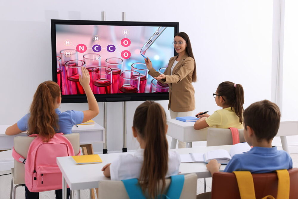 Pupils looking at a wall screen and their teacher, while the teacher uses Kramer solutions for Education