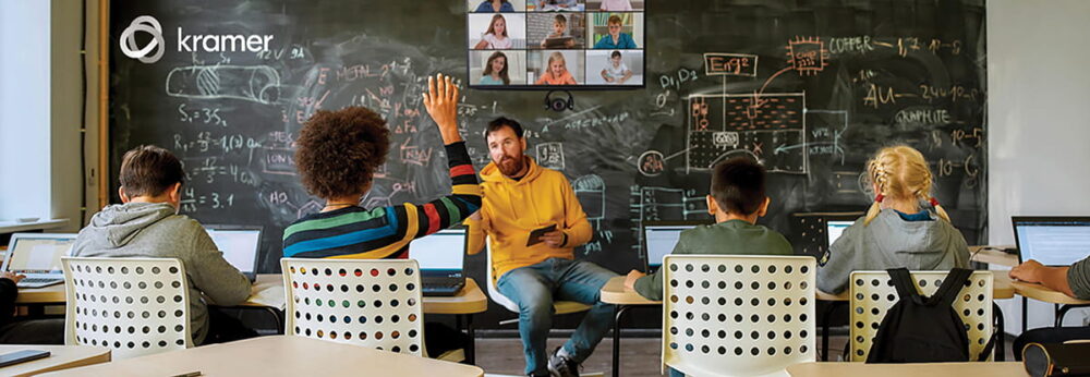 A classroom with a teacher sitting on the desk, and four students sitting, one is raising her hand