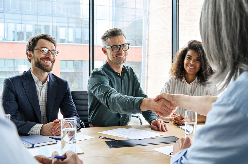 A Business meeting, three people sitting, smiling, one of them is shaking hands with another person who is sitting with his back to the camera