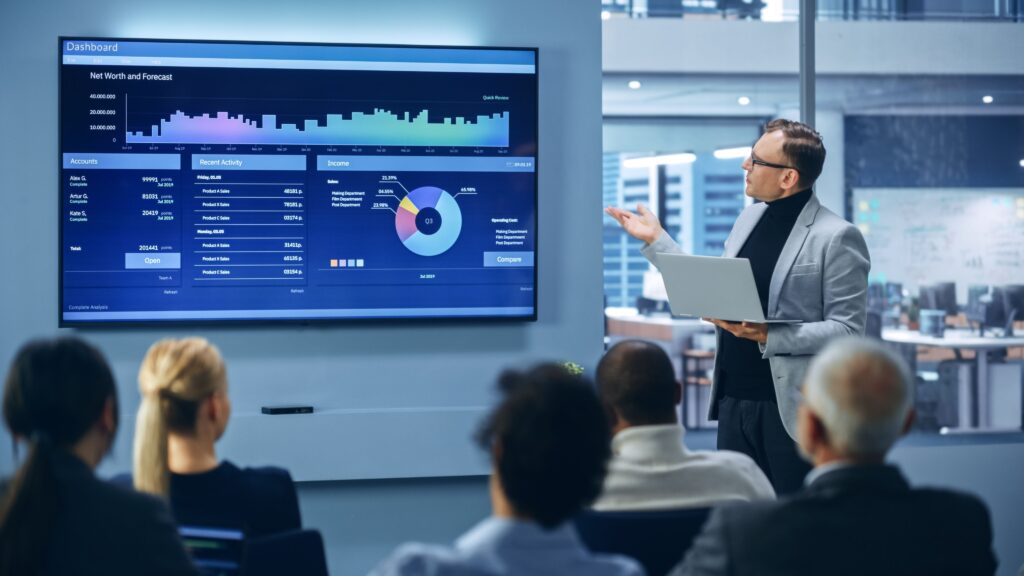 A teacher standing in front of a class, looking at a large wall screen on which we can see many charts.