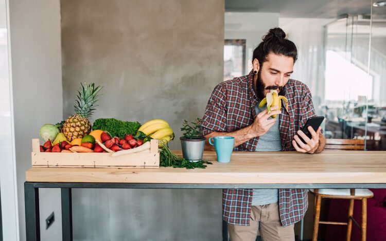 A person having a break, eating fruit and standing next to a table with fruit on it